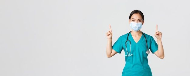 Covid-19, coronavirusziekte, concept van gezondheidswerkers. Jonge professionele aziatische vrouwelijke arts, verpleegster met medisch masker en scrubs, wijzende vingers omhoog, tonend, reclame.