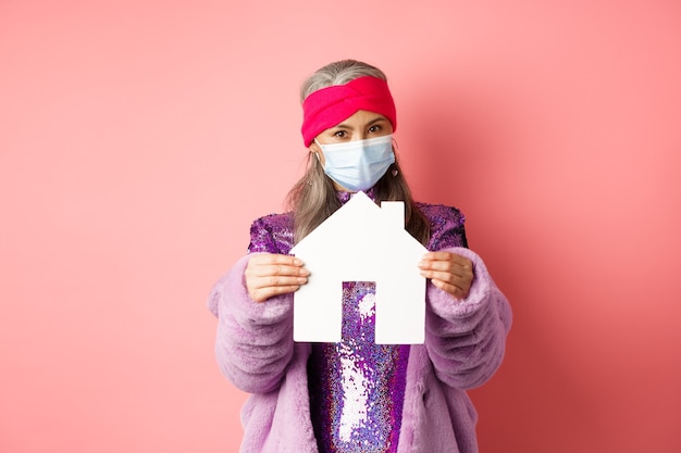 Covid-19 en vastgoedconcept. Senior aziatische vrouw in stijlvolle outfit en medisch masker met papieren huismodel, appartement zoekend, staande over roze achtergrond.