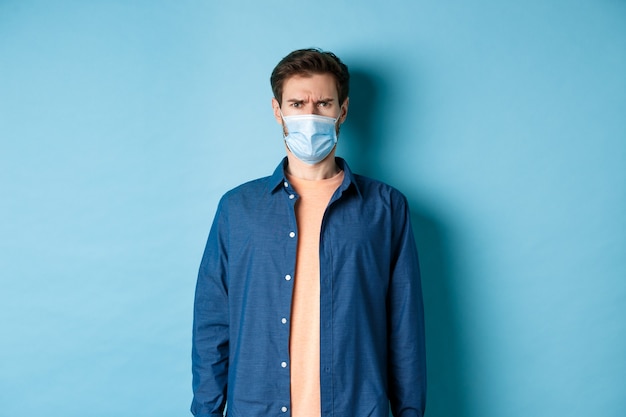 Covid-19 en zorgconcept. Teleurgestelde man in medisch masker fronsen, op zoek verward, staande op een blauwe achtergrond.