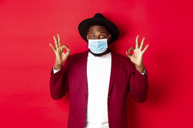 Covid-19 and fashion concept. Stylish african american man in hat and blazer, wearing face mask and showing okay sign, standing over red background