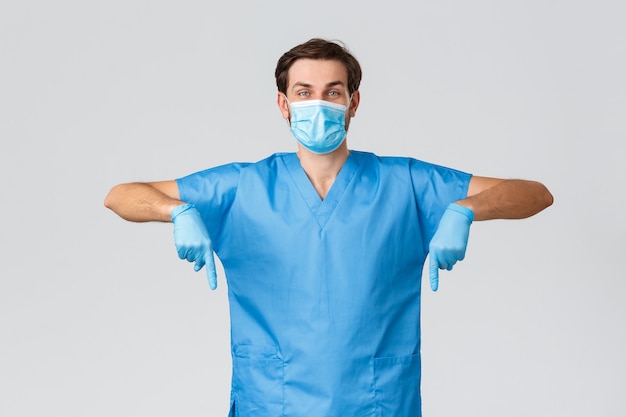 Covid-19, gezondheidswerkers en ziekenhuisconcept. Vrolijke enthousiaste arts in scrubs, vecht tegen coronavirus, draag beschermende uitrusting, medisch masker en handschoenen, wijzend naar beneden om promo te tonen