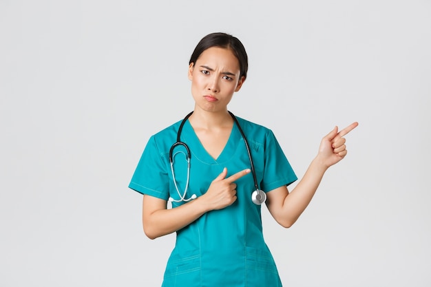 Covid-19, gezondheidswerkers, pandemisch concept. Teleurgesteld en boos Aziatische vrouwelijke therapeut, verpleegster in scrubs boos camera kijken, mokkend en wijzend rechterbovenhoek, uitleg wachten.