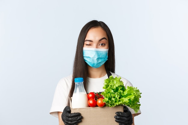 Covid-19, grocery store, employment, small business and preventing virus concept. Smiling asian female shop manager, cashier in medical mask and gloves, taking care of your food order.