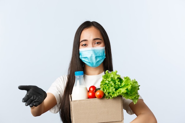 Covid-19, grocery store, employment, small business and virus concept. Cute young asian shop employee, cashier in medical mask and gloves waiting for money for order, holding packed groceries.