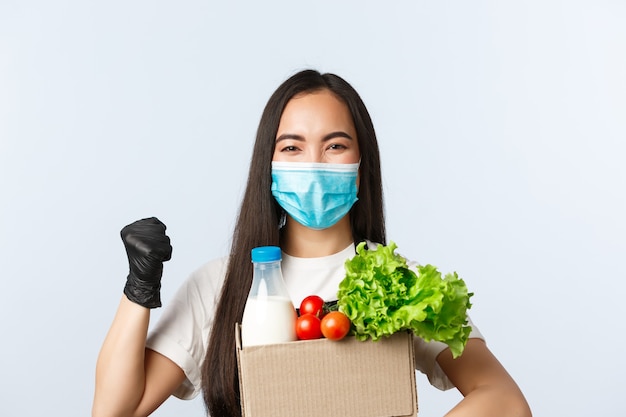 Covid-19, grocery store, employment, small business and virus concept. Enthusiastic asian female employee, shop cashier in medical mask, fist pump encourage buy safe, giving bag with groceries