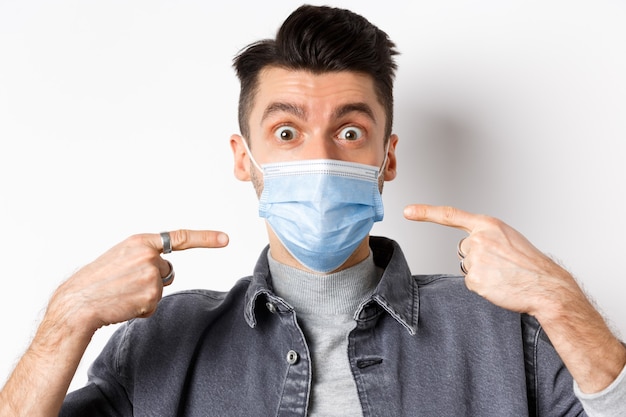 Covid-19, health and lifestyle concept. Excited guy pointing fingers at his sterile medical mask, use preventive measures from catching coronavirus on quarantine, white background.