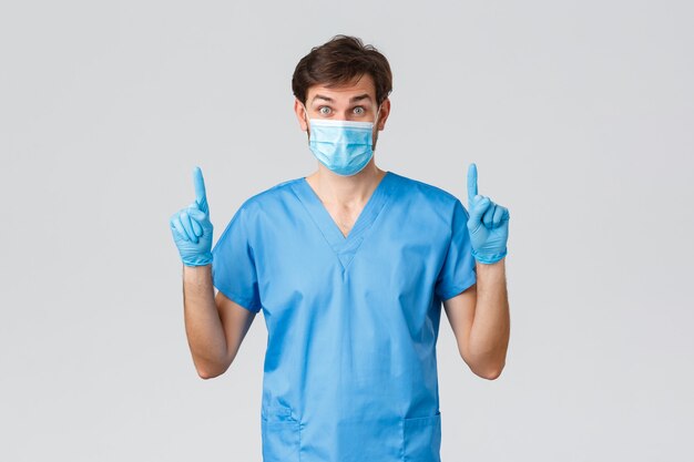 Covid-19, healthcare workers and hospital concept. Surprised and excited doctor in medical mask, rubber gloves and scrubs, hearing interesting news, pointing advertisement up, top promo