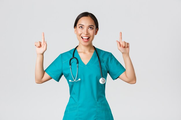 Covid-19, healthcare workers, pandemic concept. Amused cheerful asian female nurse, doctor or surgeon in scrubs pointing fingers and looking up with satisfied face, standing white background.