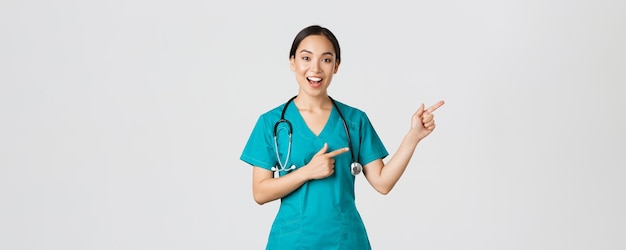 Covid-19, healthcare workers, pandemic concept. Glad smiling, beautiful asian doctor, female therapist in scrubs or nurse pointing fingers upper right corner, showing way to patient, white background.