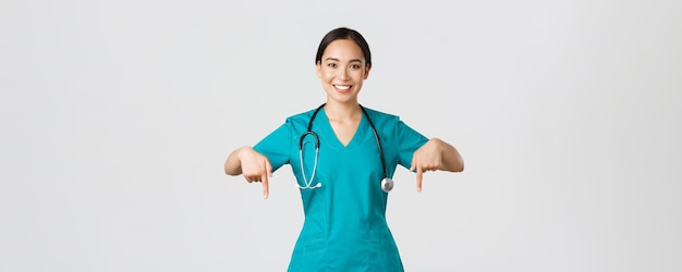 Covid-19, healthcare workers, pandemic concept. Smiling pleasant asian female doctor, therapist or physician in scrubs with stethoscope, pointing fingers down, show clinic banner