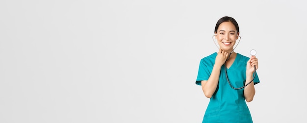 Covid-19, healthcare workers and preventing virus concept. Cheerful smiling asian female physician, doctor consult patient, examine person with stethoscope, standing white background