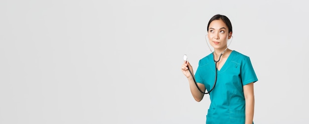 Covid-19, healthcare workers and preventing virus concept. Young focused asian female doctor, physician in scrubs, examine patient lungs, using stethoscope, standing thoughtful white background