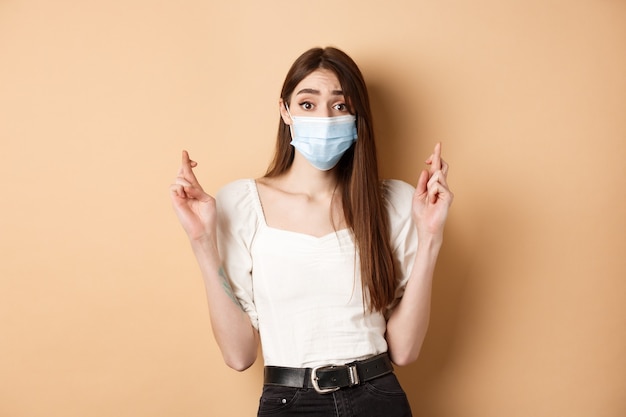 Covid-19 and lifestyle concept. Hopeful girl in face mask praying with fingers crossed, looking with hope at camera, standing on beige background.