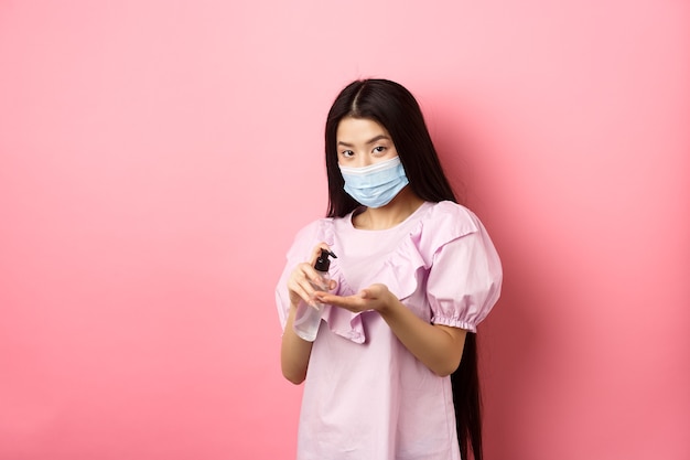 Covid-19, lifestyle and health concept. Beautiful asian woman in medical mask clean hands with sanitizer, apply antiseptic on palms, standing on pink background.