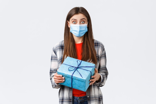 Covid-19, lifestyle, holidays and celebration concept. Excited cute birthday girl holding wrapped box, curious what inside, receive gift, wearing medical mask to prevent coronavirus outbreak