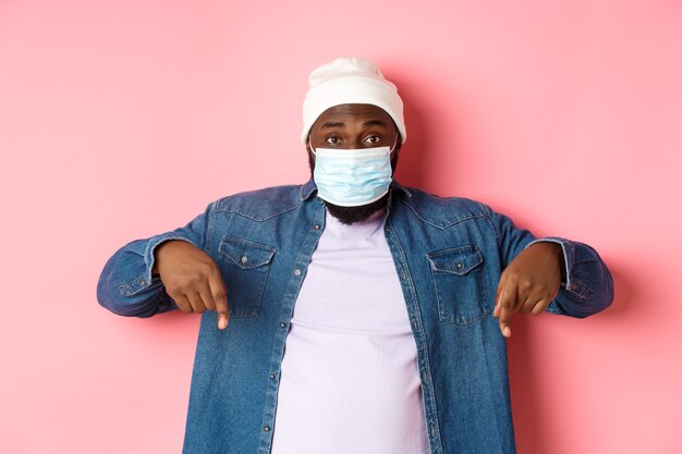 Covid-19, lifestyle and lockdown concept. Handsome african-american man in face mask, pointing fingers down, showing promo offer on bottom, standing over pink background