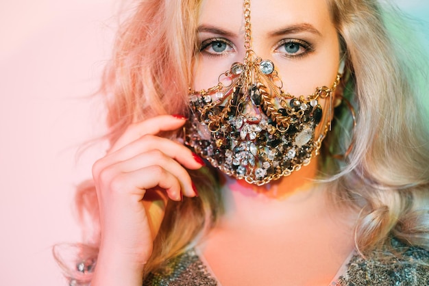 Covid-19 mode. Quarantaine accessoire. Feestelijke uitstraling. Portret van sensuele blonde vrouw in luxe gouden sieraden ketting gezichtsmasker op pastel blauw roze neon licht achtergrond.