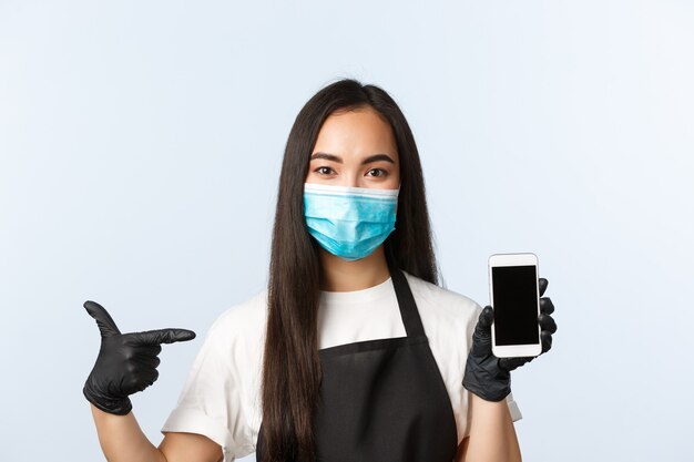 Covid-19 pandemic, coffee shop, small business and preventing virus concept. Attractive asian female employee, cafe worker in medical mask and gloves pointing at mobile phone screen