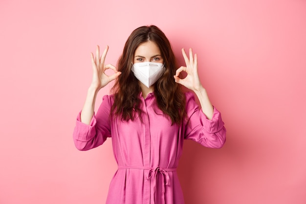 Covid-19, pandemic and lifestyle concept. Cheerful girl shows okay sign, wears medical respirator as preventive measure from corona, pink wall.