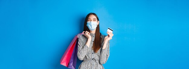 Covid-19 pandemic and lifestyle concept happy girl wear face mask on shopping showing plastic person
