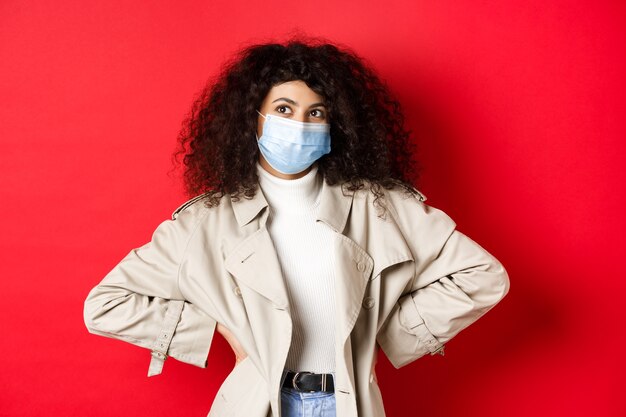 Covid-19, pandemic and quarantine concept. Stylish enthusiastic woman in medical mask and trench coat, smiling and looking at upper left corner, red wall.