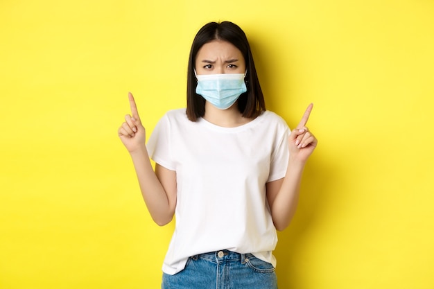 Covid-19, концепция пандемии и социального дистанцирования. Разочарованная азиатская девушка в медицинской маске, расстроенная хмурится и указывая пальцами на логотип, стоя на желтом фоне.