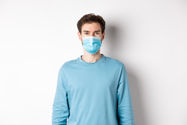 Covid-19, pandemic and social distancing concept. Young man wearing medical mask from coronavirus, standing over white background