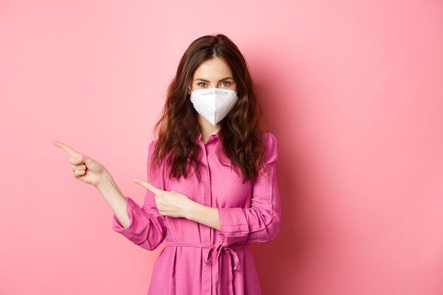 Covid-19, pandemie en levensstijlconcept. stijlvol modern meisje in gezichtsmasker, wijzende vingers naar links, met copyspace opzij, staande tegen roze muur.