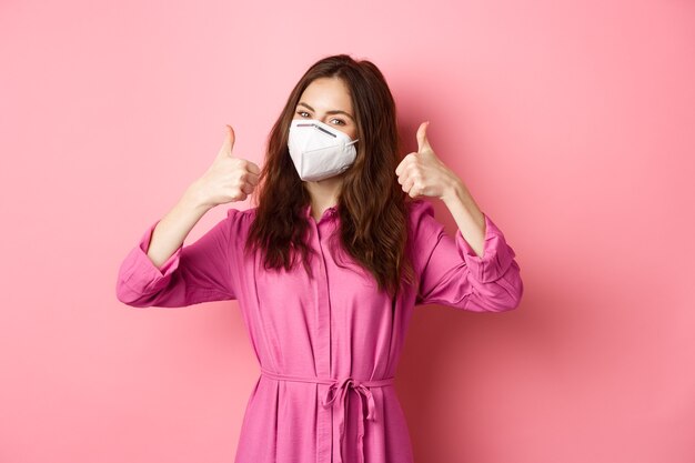 Covid-19, pandemie en levensstijlconcept. vrolijk meisje verschijnt duimen, draagt medische gasmasker als preventieve maatregel tegen corona, roze muur.