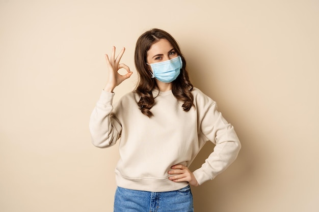 Covid-19, pandemie en quarantaineconcept. Jonge vrouw draagt een medisch gezichtsmasker tijdens de uitbraak van coronavirus-omicron, vertoont een goed teken en staat op een beige achtergrond