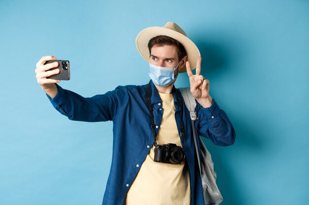 Covid-19, pandemie en reisconcept. Gelukkig en positieve man in toeristische hoed selfie te nemen en vredesteken te tonen, poseren in de buurt van bezienswaardigheden, blauwe achtergrond.