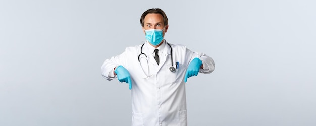 Covid-19, preventie van virussen, gezondheidswerkers en vaccinatieconcept. Geschokt hijgende mannelijke arts met medisch masker en kruidnagel reageert op het uitbraakpercentage van het coronavirus en wijst met de vingers naar beneden