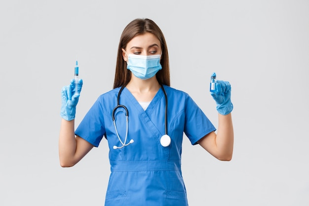 Covid-19, preventing virus, health, healthcare workers and quarantine concept. Female nurse or doctor in blue scrubs, medical mask and gloves, look at ampoule with coronavirus vaccine, hold syringe