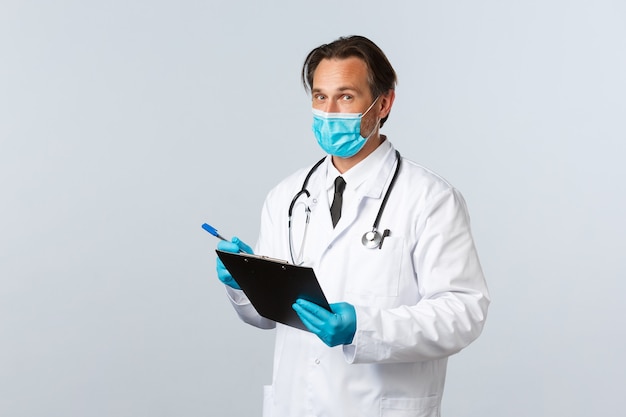 Covid-19, preventing virus, healthcare workers and vaccination concept. Professional doctor in medical mask and gloves, holding clipboard, writing down patient symptoms, fight coronavirus in clinic