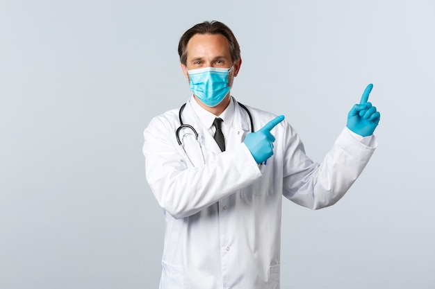 Covid-19、ウイルス、医療従事者、予防接種の概念を防ぎます。白衣、医療マスク、右上の広告、白い背景を指す手袋でフレンドリーな医師の笑顔