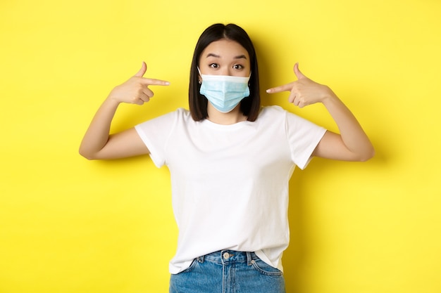 Covid-19, quarantaine en social distancing concept. Jonge aziatische vrouw in wit t-shirt, wijzend op haar medische masker van coronavirus, gele achtergrond