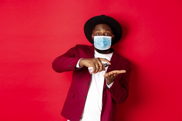 Covid-19, quarantine and holidays concept. Handsome Black man in face mask and party outfit, disinfecting hands with hand sanitizer, using antiseptic, standing over red background.