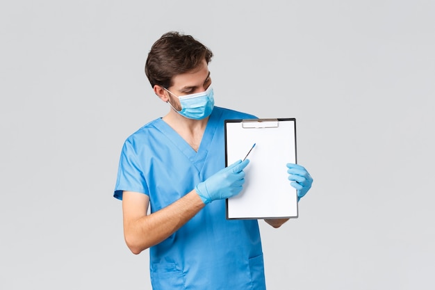 Covid-19, quarantine, hospitals and healthcare workers concept. Physician explain important of taking measures to protect from coronavirus. Doctor in scrubs and medical mask pointing at clipboard.