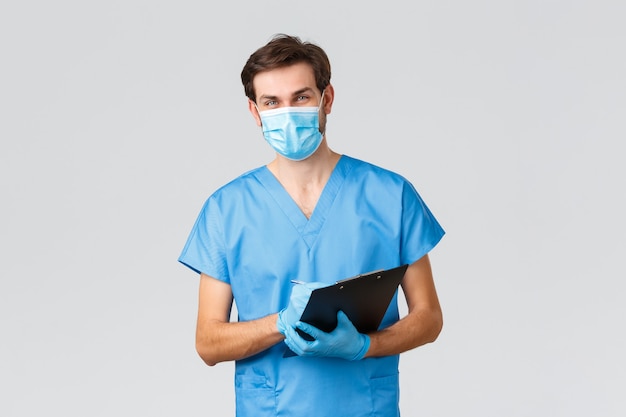 Covid-19, quarantine, hospitals and healthcare workers concept. Smiling young doctor, nurse in medical mask, scrubs and gloves, writing down patient symptoms or info at clipboard, fill in blank in ER