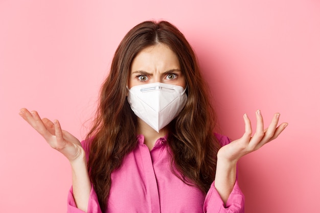 Covid-19, quarantine and social distancing concept. Close up portrait of confused brunette woman in respirator shrugging shoulders, look puzzled and upset, standing over pink wall.