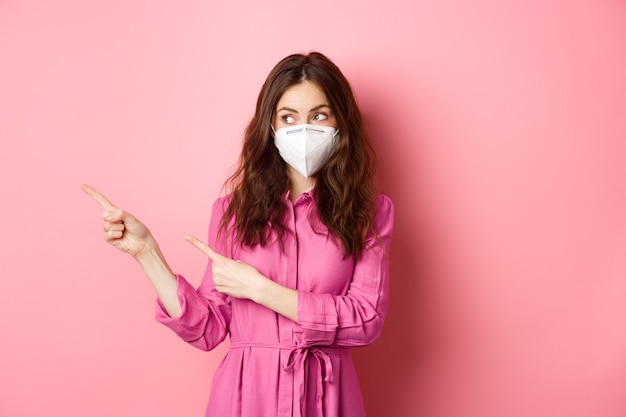 Covid-19, quarantine and social distancing concept. Curious stylish woman in respirator, looking and pointing fingers aside, showing promo text logo, standing over pink wall.