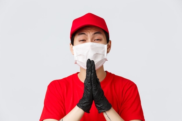 Covid-19、自己検疫のオンラインショッピングと配達のコンセプト。医療用マスクと手袋のしがみつくアジアの宅配便、物乞い、物乞い、助けが必要、灰色の背景に立っている