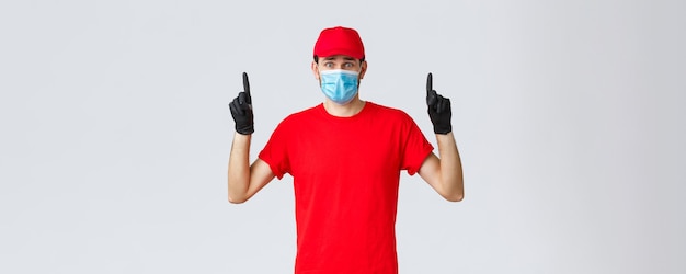 Covid-19, self-quarantine, online shopping and shipping concept. Nervous or scared, worried delivery man in red uniform cap, t-shirt, frowning pointing fingers up at bad news, wear mask and gloves