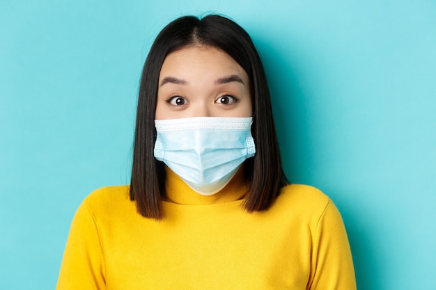 Covid-19, sociaal distantiëren en pandemisch concept. Close-up van verrast Aziatische vrouw in medisch masker, wenkbrauwen optrekken en kijk verbaasd naar camera, blauwe achtergrond.