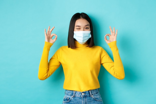 Covid-19, social distancing and pandemic concept. Cheerful asian girl in medical mask showing okay gestures, winking at camera, showing approval, standing over blue background.