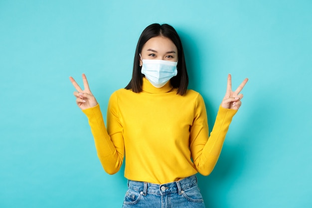 Covid-19, social distancing and pandemic concept. Kawaii asian woman showing peace signs and smiling happy, wearing medical mask from coronavirus disease, standing over blue background.