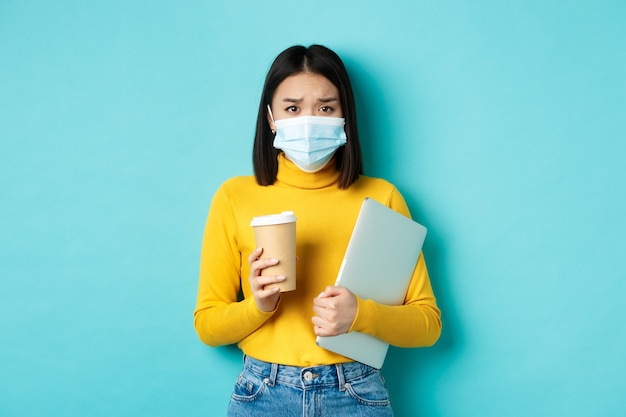 Covid-19, social distancing and pandemic concept. Worried asian woman in medical mask, frowning sad, holding laptop for work and cup of coffee