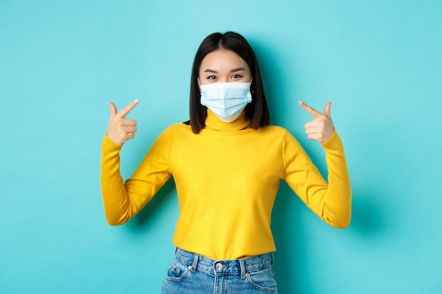 코비드-19, 사회적 거리 및 전염병 개념. 젊은 아시아 여성은 코로나바이러스로부터 자신을 보호하고 의료 마스크를 손가락으로 가리키며 파란색 배경 위에 서 있습니다
