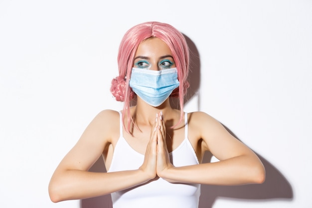 Covid-19, social distancing and people concept. Close-up of cute party girl in pink wig and medical mask, holding hands together and looking left, begging for help