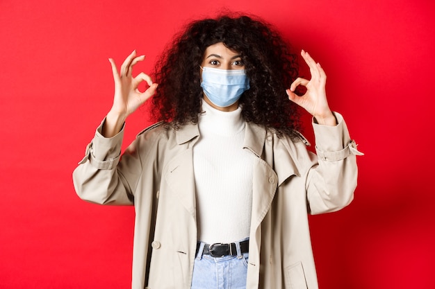 Covid-19, social distancing and quarantine concept. Fashionable woman with curly hair, wearing medical mask and trench coat, showing okay gestures, red background.